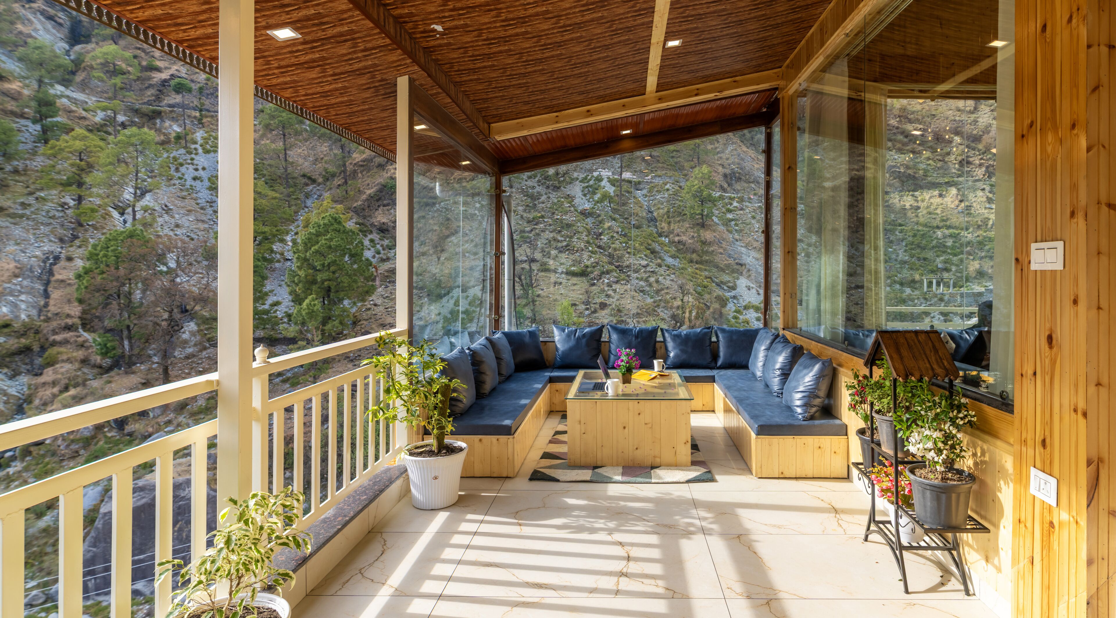 Une terrasse en bois dotée d’un espace salon intégré, de grandes fenêtres offrant une vue sur un paysage montagneux et de plantes en pot.