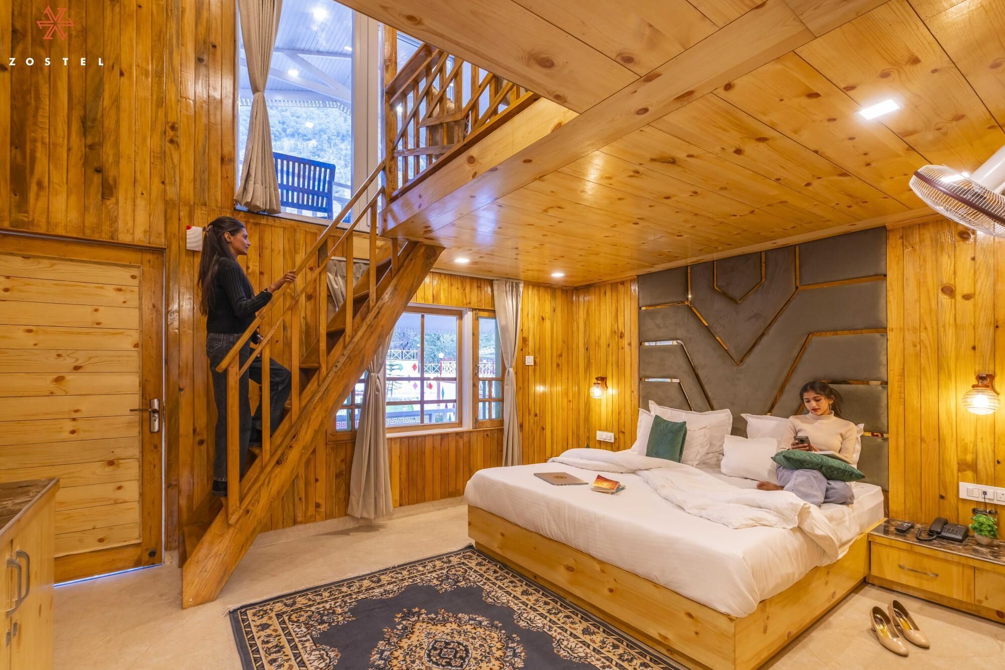 A wooden cabin interior with a bed, a staircase, and a view of the outside.