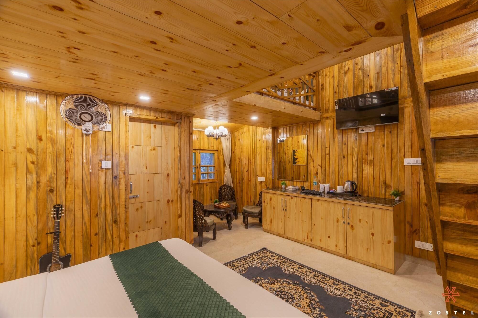 A wooden interior with a bed, a fan, a guitar, a TV, and a kitchen area.