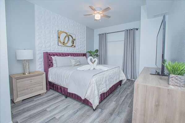 A bedroom with a bed, a nightstand, a ceiling fan, a window with curtains, and a plant on a wooden table.