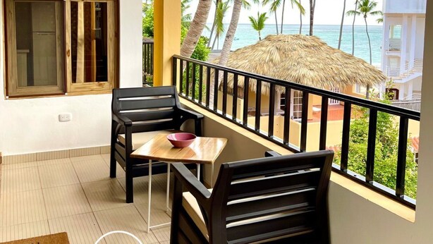 A balcony with a wooden table and chairs, a thatched roof, and a view of palm trees and the ocean. - C-402 Loft apt in Punta Cana, los Corales Beach/pool (Punta Cana)