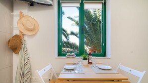 Un comedor con una mesa de madera dispuesta para dos, una ventana con contraventanas verdes y un sombrero y una cesta colgados en la pared.