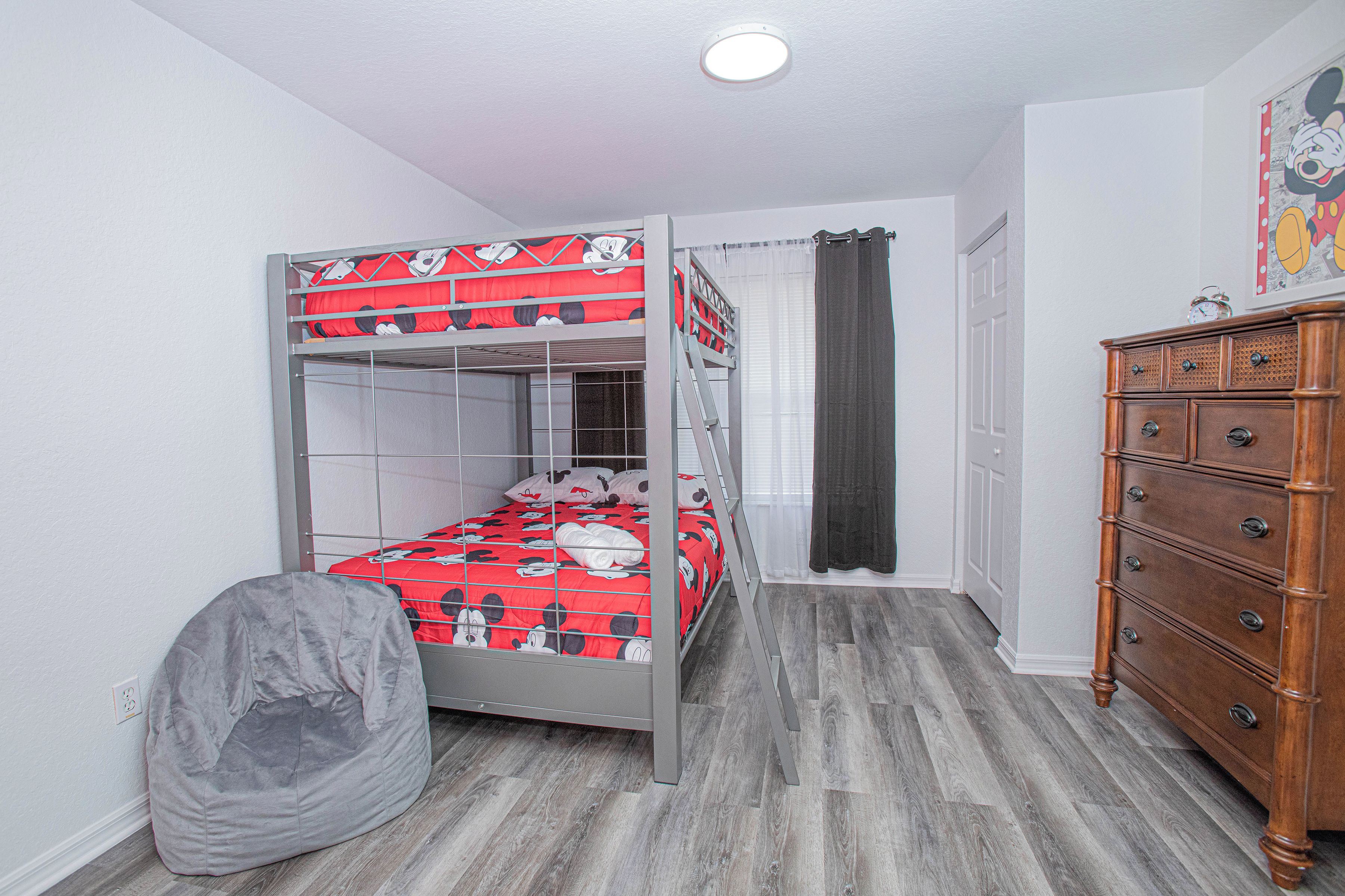A bunk bed with Mickey Mouse bedding, a wooden dresser, and a grey bean bag chair in a room with wooden flooring and white walls.