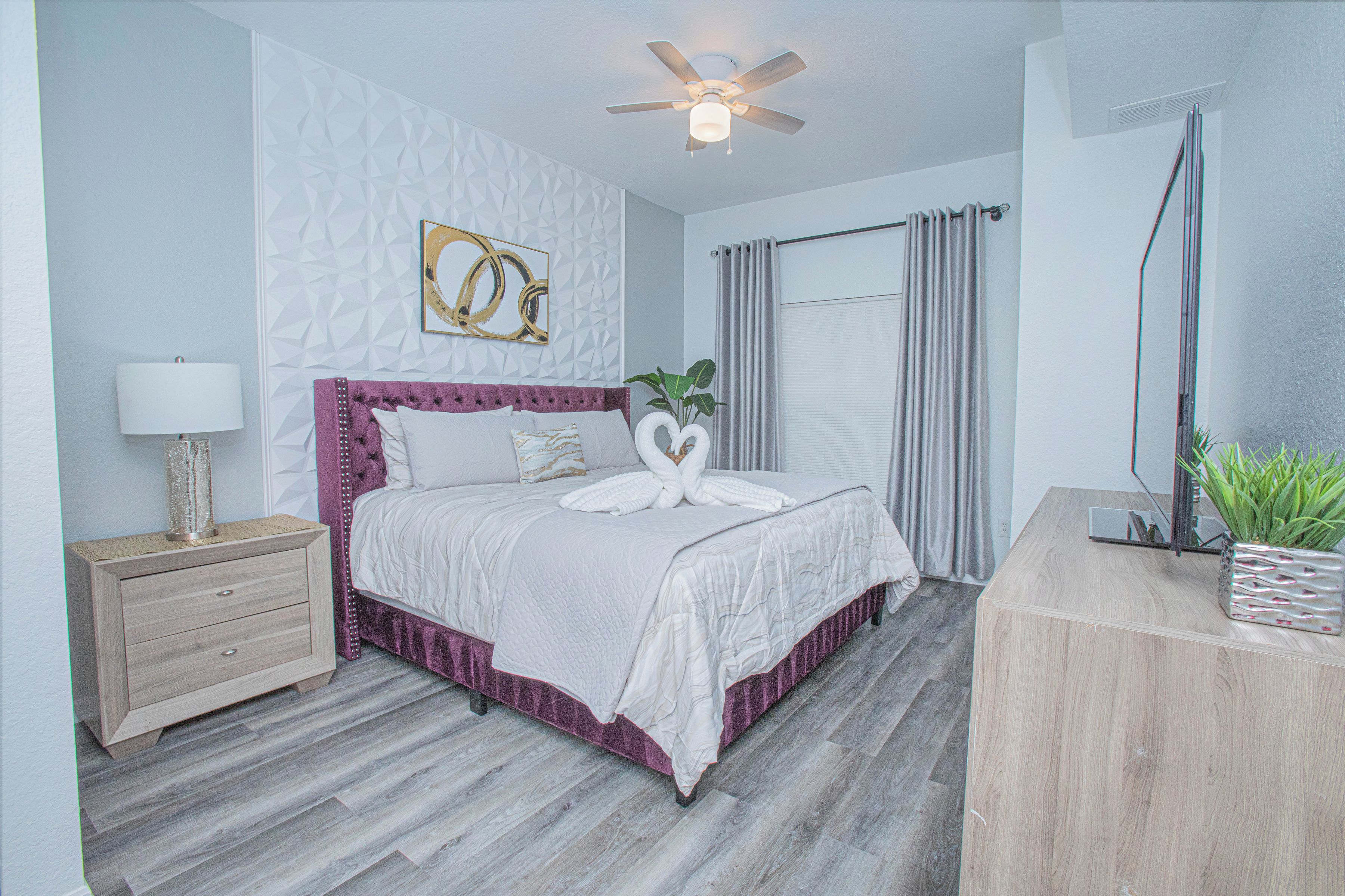 A bedroom with a bed, a nightstand, a ceiling fan, a wall-mounted artwork, and a potted plant.