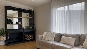 Un soggiorno con un divano beige, un mobile porta TV nero con piante e un'ampia finestra con tende leggere.