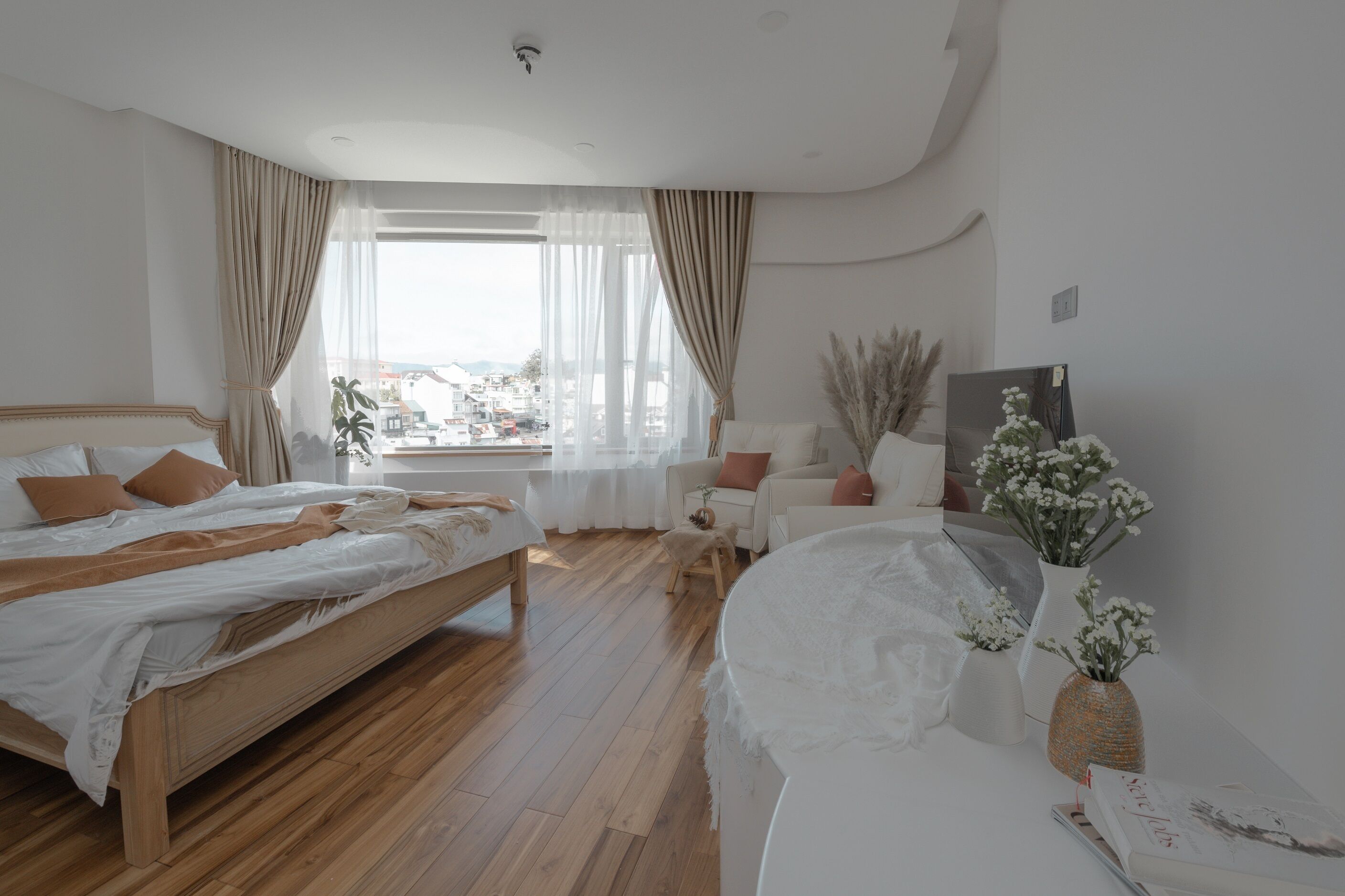 Ein modernes Schlafzimmer mit einem hölzernen Bett, einem weißen Sofa und Blick auf die Stadt durch transparente Vorhänge.
