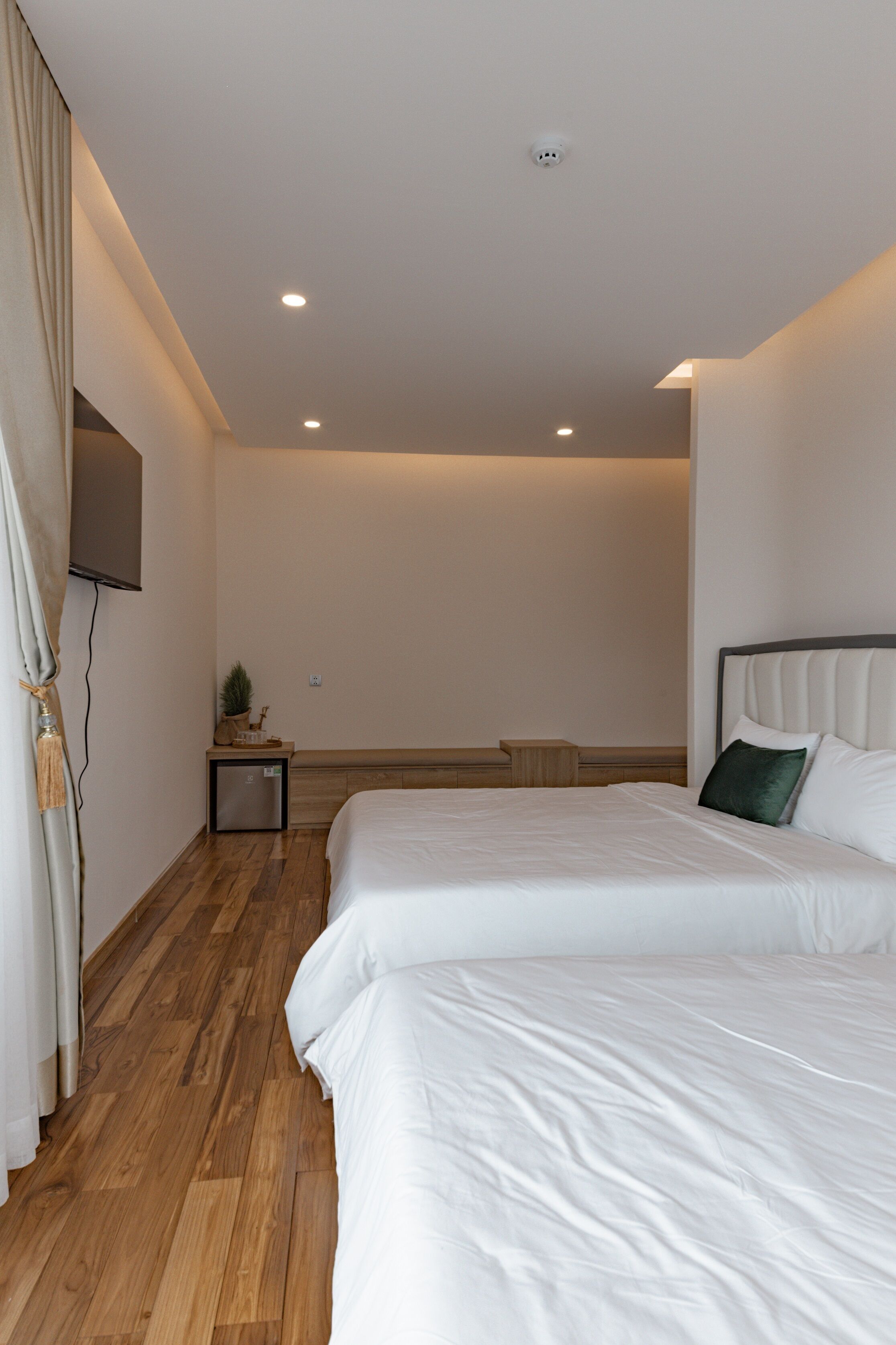 A modern hotel room with two beds, wooden flooring, a small bedside table, a TV mounted on the wall, and a ceiling light.