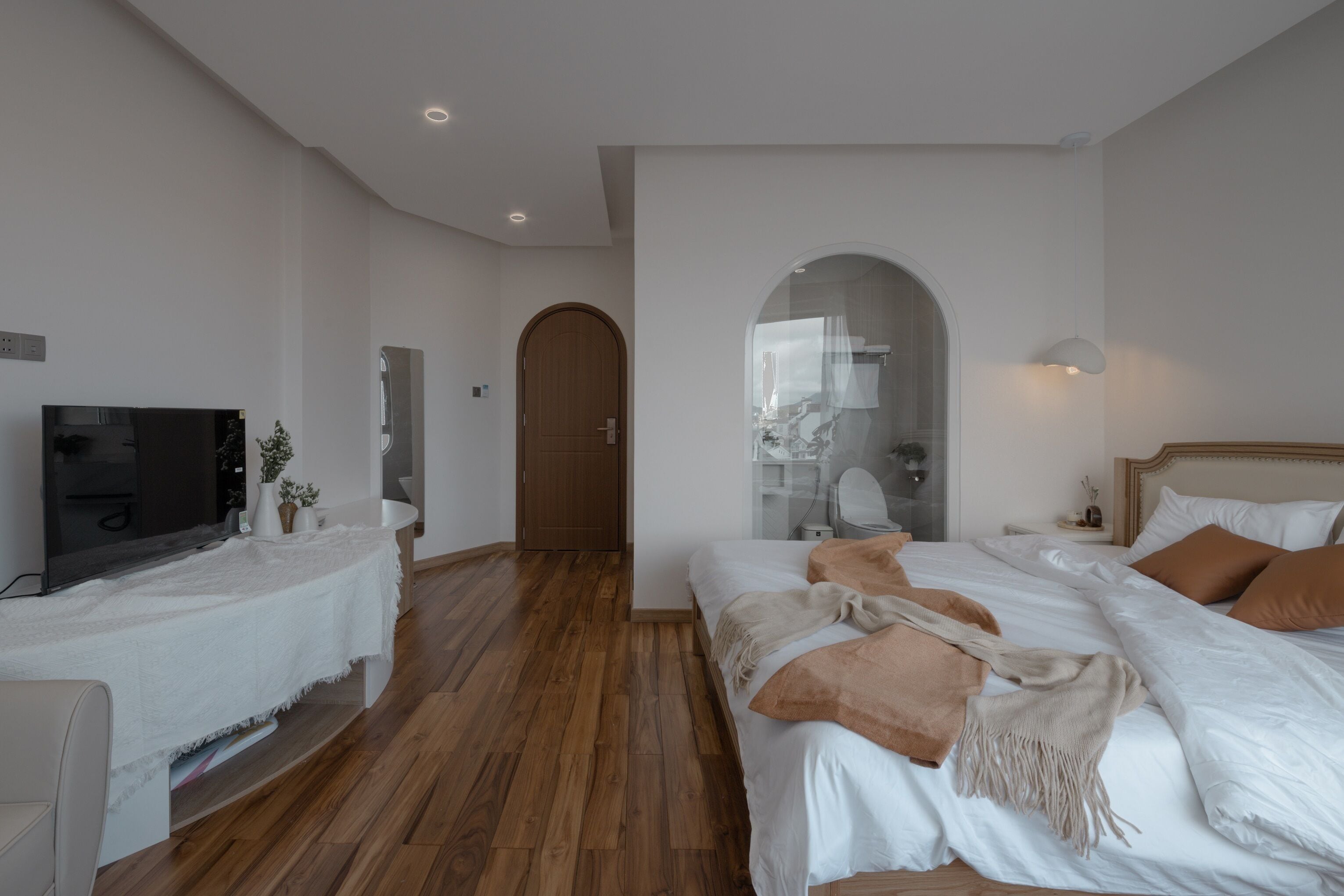 Ein Schlafzimmer mit Holzboden, einem Bett mit weißer Bettwäsche und braunen Kissen, einem Fernseher auf einem weißen Tisch und einem Spiegel an der Wand.
