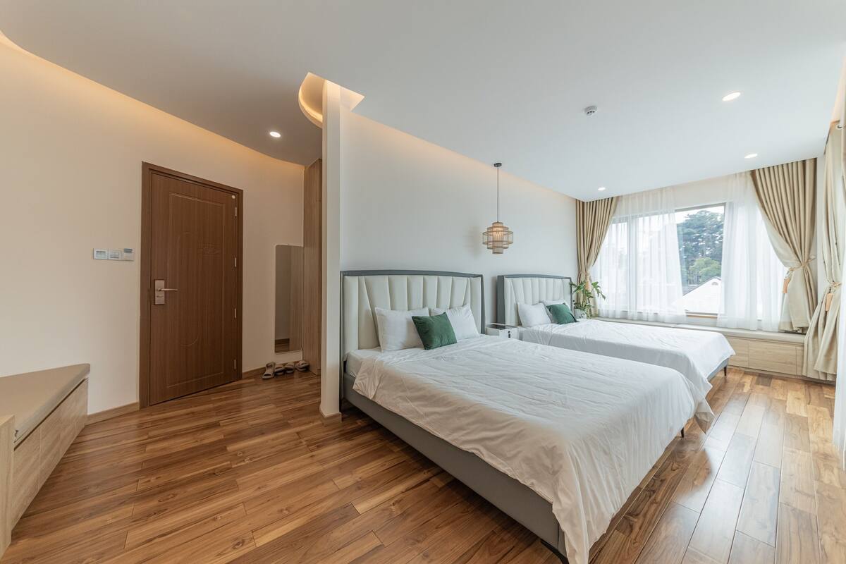 Ein modernes Schlafzimmer mit zwei Betten, Holzboden, einem großen Fenster mit Vorhängen und einer Pendelleuchte.
