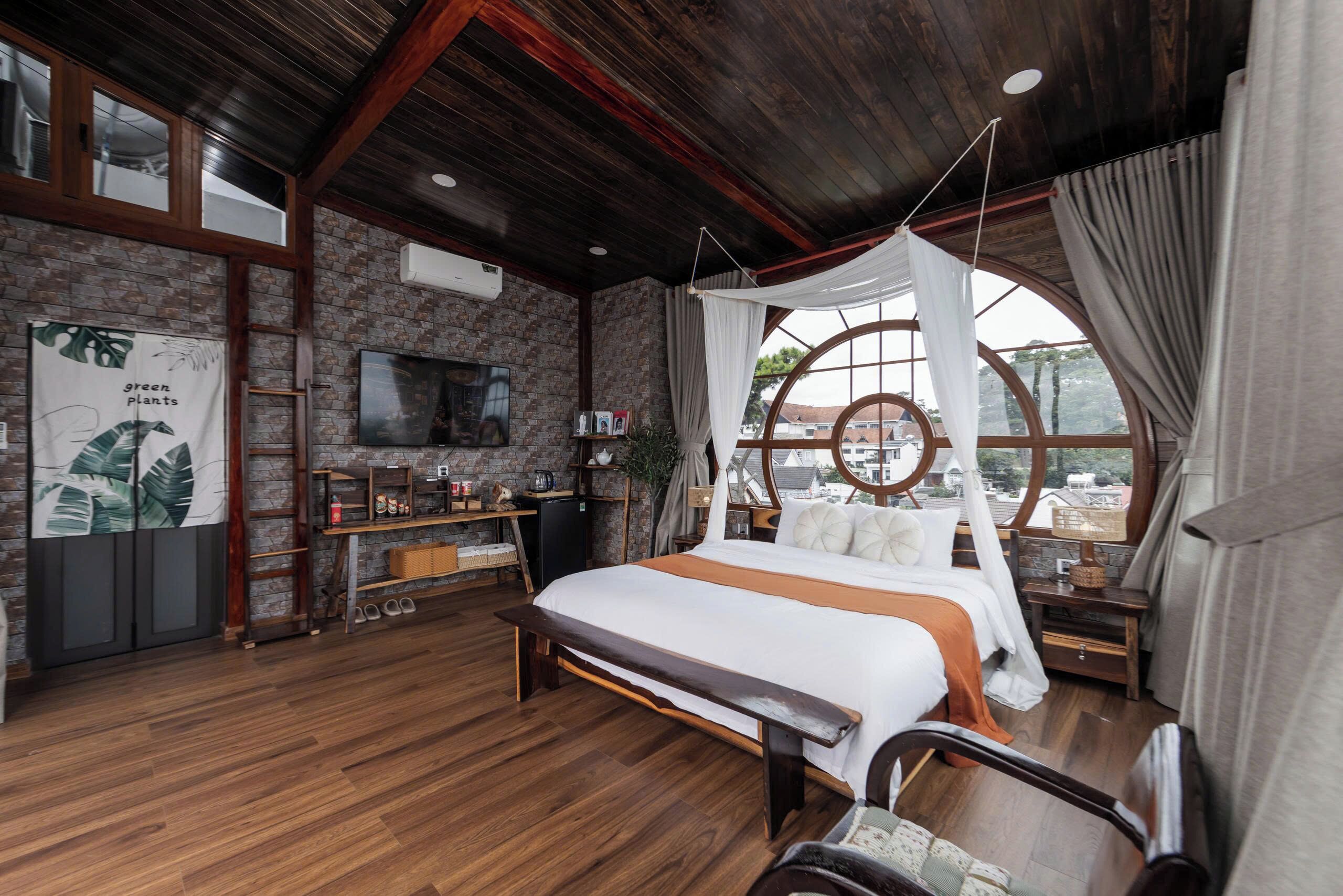 A bedroom with a large bed, wooden floor, a TV, a window with curtains, and a brick wall.
