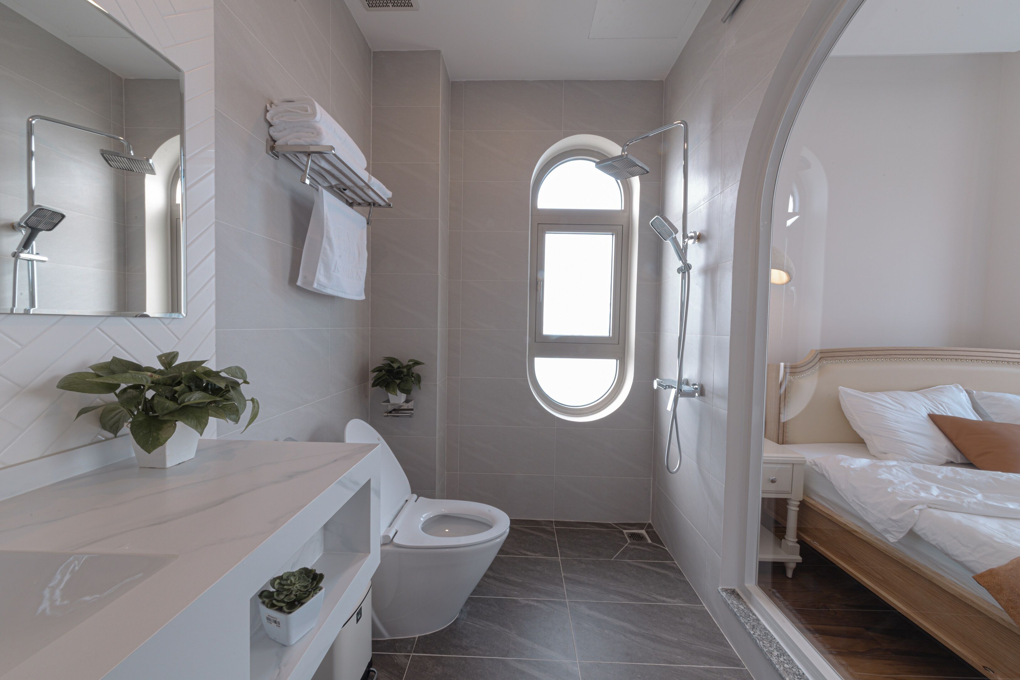 A modern bathroom with a shower, toilet, and sink. There is a window providing natural light and a bed with a headboard in the adjacent bedroom.