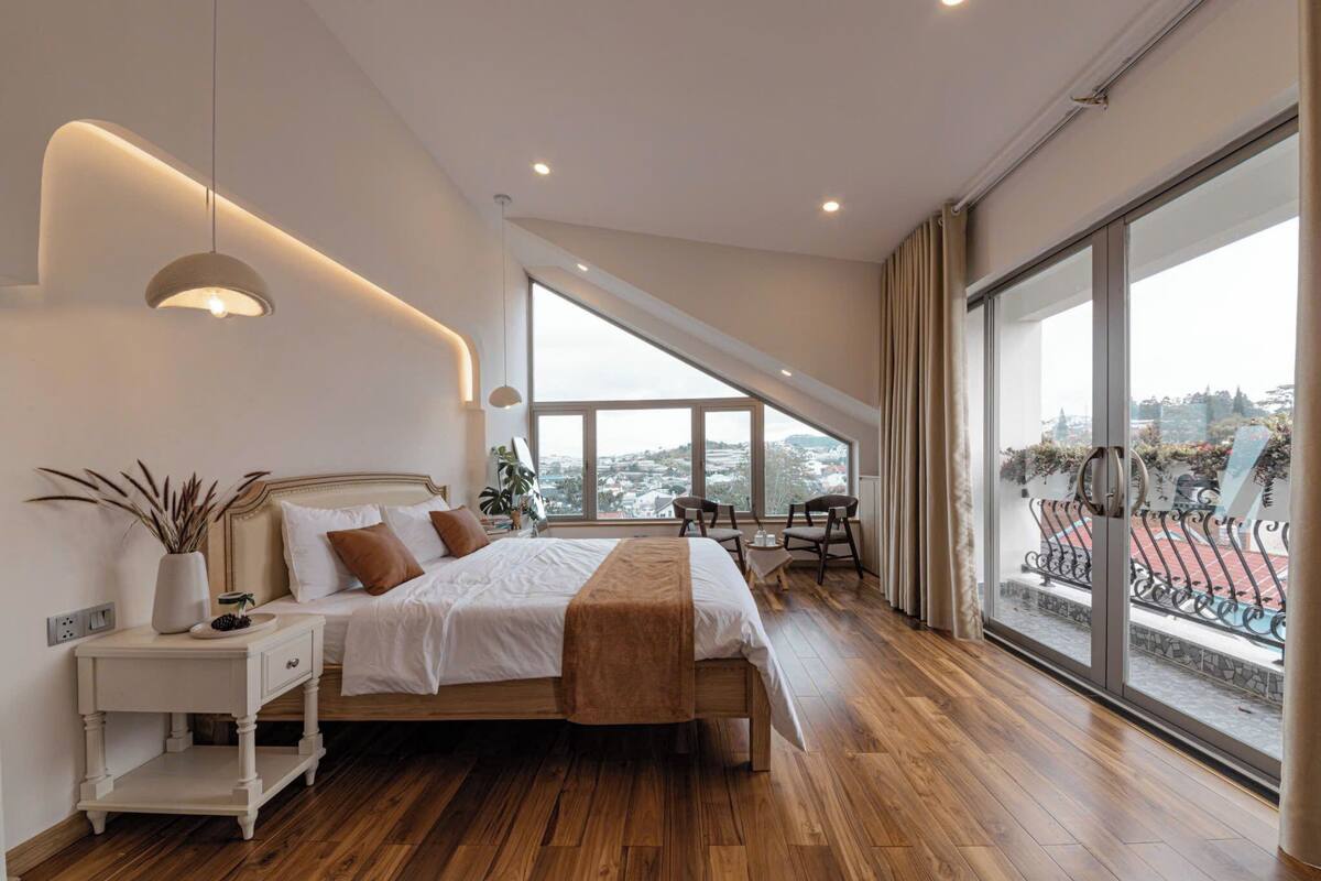 Ein modernes Schlafzimmer mit einem großen Bett, Nachttischen, einem kleinen Tisch daneben, einem Fenster mit Blick auf die Stadt und einem Balkon mit Gartenmöbeln.