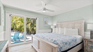 Un dormitorio con cama, ventilador de techo, vista a una piscina y palmeras, y una mesita de noche de madera con lámpara.