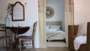 A cozy bedroom with a bed, a chair, a table with a vase and mugs, and a decorative wall piece.