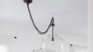 Une table à manger blanche, surmontée d’un luminaire suspendu en verre transparent, un vase blanc contenant des feuilles vertes et un plateau en bois.