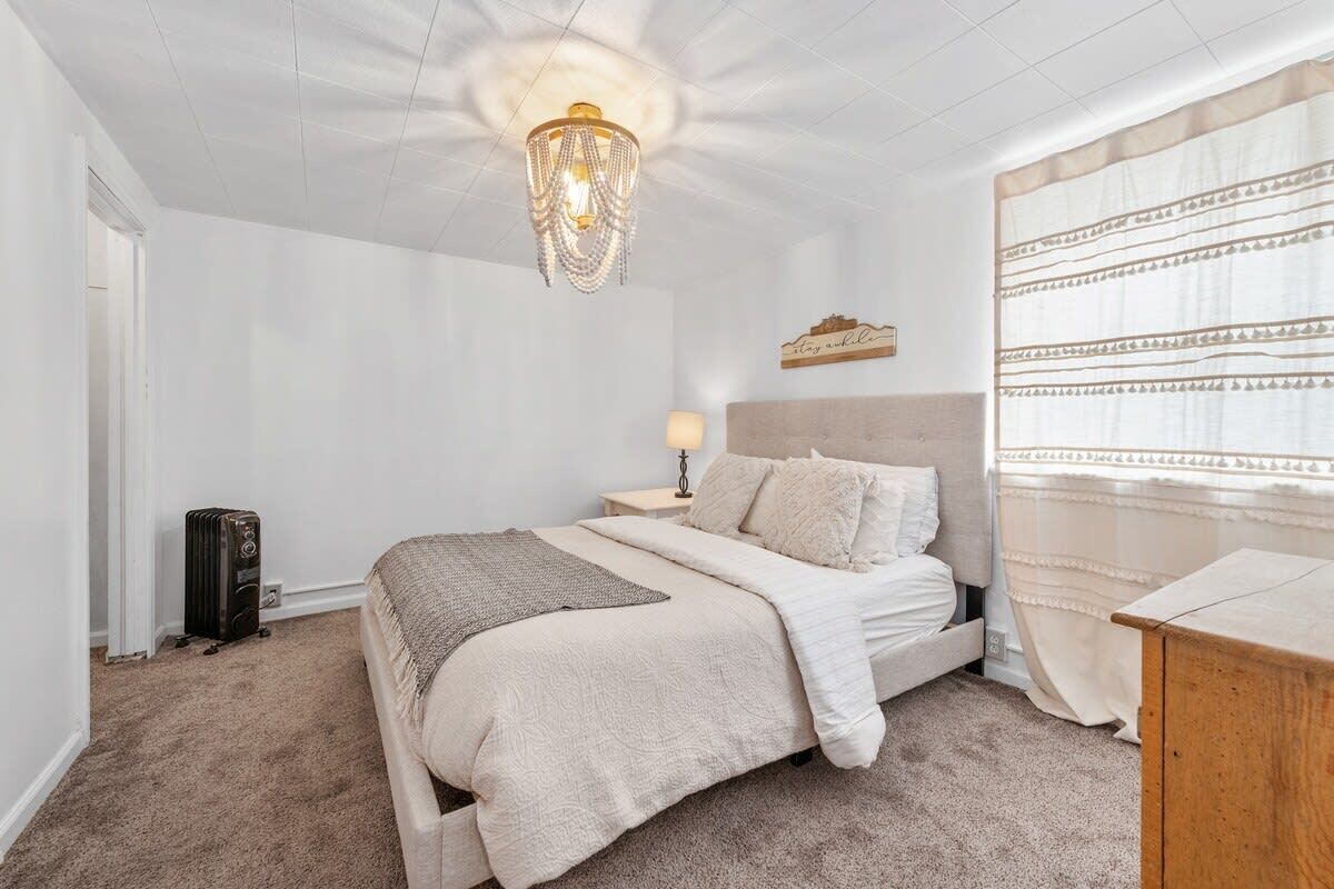A bedroom with a bed, a nightstand, a lamp, a chandelier, and a window with curtains.