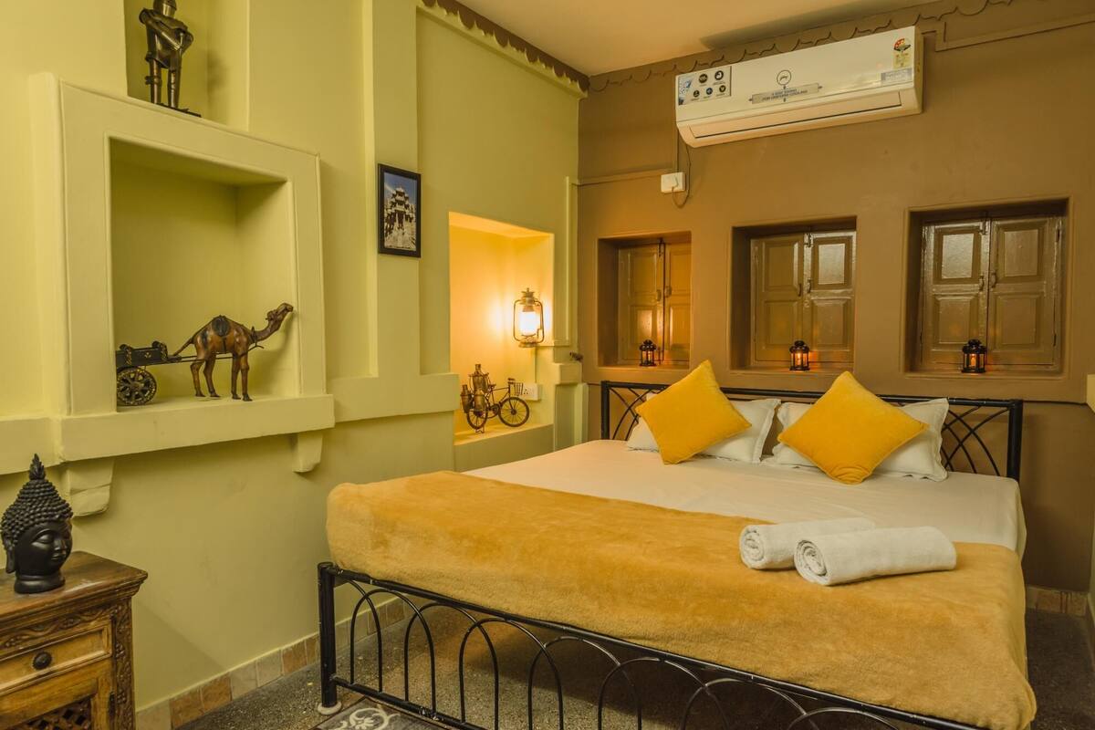 Una habitación de hotel con una cama, un estante con una escultura de camello y una bicicleta, un aire acondicionado instalado en la pared y una ventana con vistas.