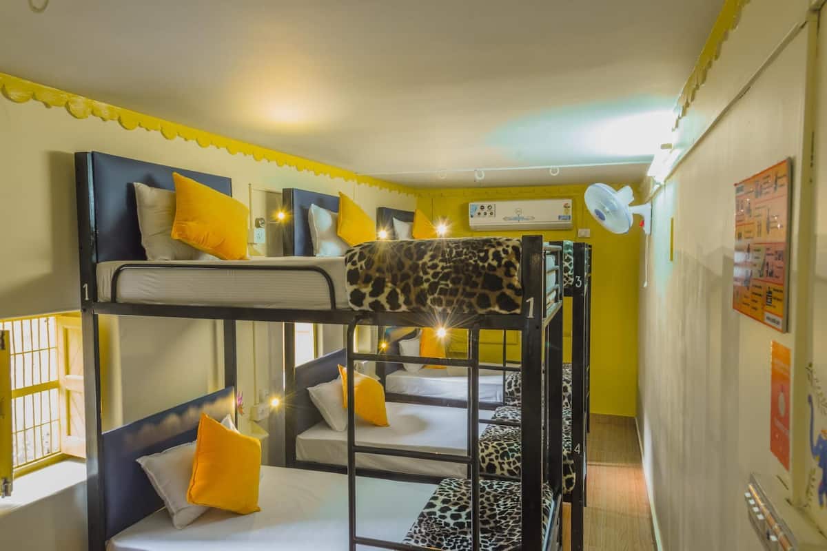 Habitación con literas, ropa de cama amarilla y blanca, ventilador de techo y una ventana con barrotes.