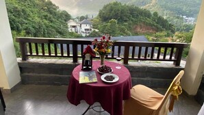 A balcony with a table set for two, a vase with flowers, and a book. The balcony overlooks a mountainous landscape with a house in the background.