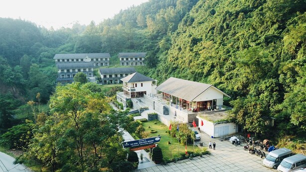 A resort with multiple buildings surrounded by lush greenery, a signboard, and a parking area with vehicles.