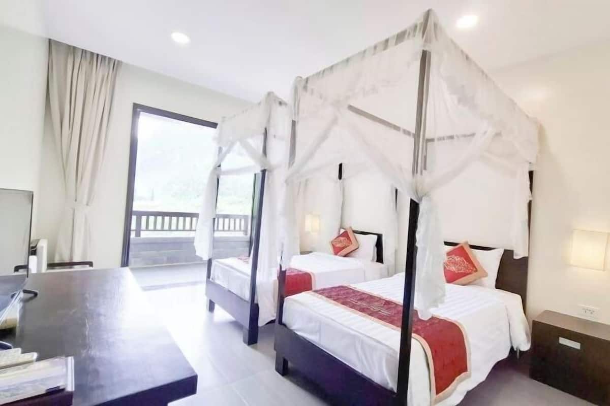 Une chambre avec deux lits, chacun doté d’un baldaquin, de draps blancs et d’éléments décoratifs rouges. On y trouve un bureau avec une chaise, une fenêtre avec des rideaux et une vue sur l’extérieur.