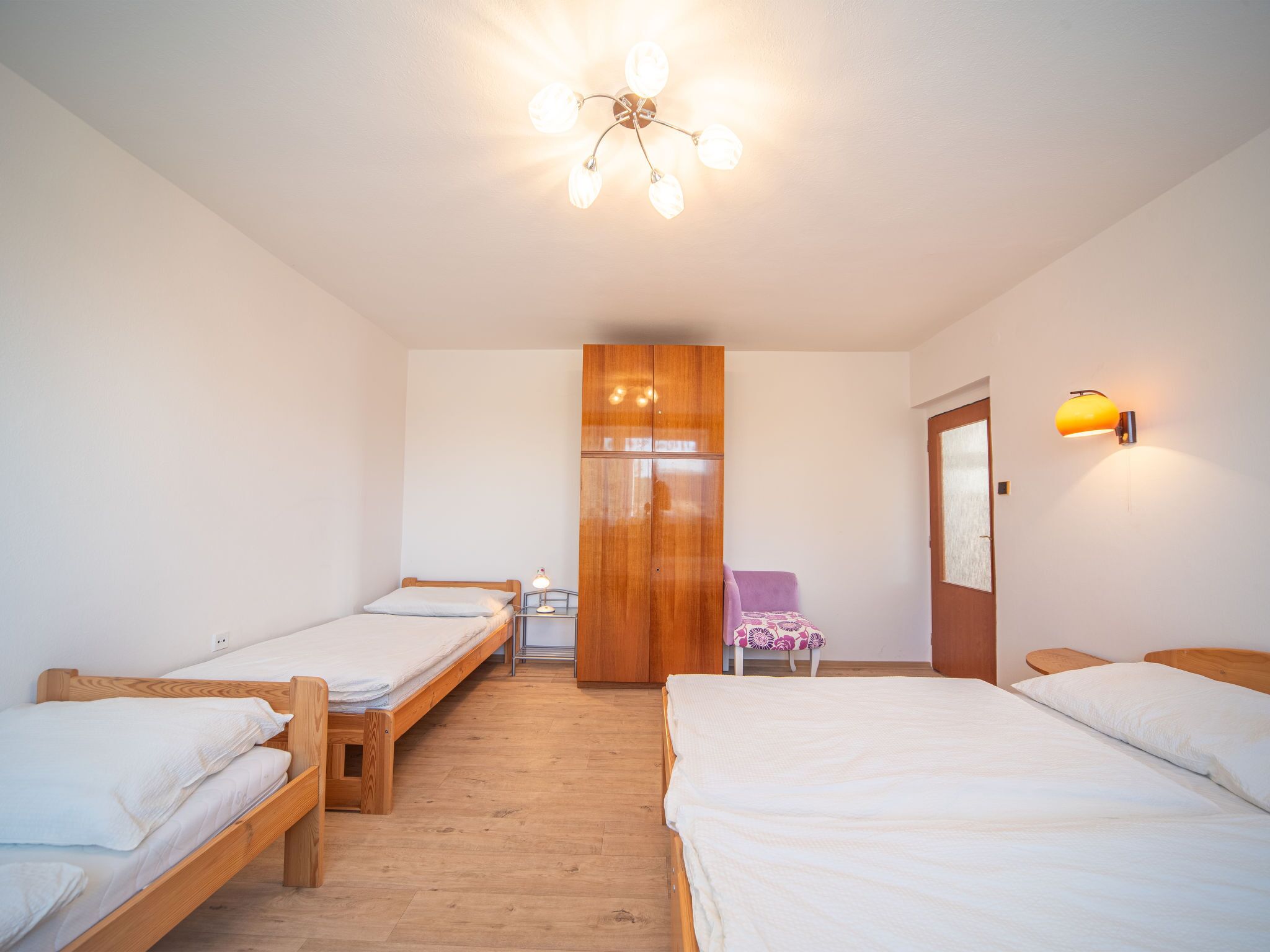 A room with two single beds, a wooden wardrobe, a purple chair, and a wall-mounted lamp.