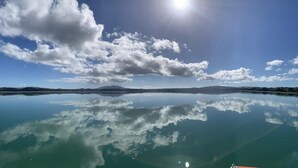 Ein ruhiger See mit klaren Spiegelungen des Himmels und der Wolken, umgeben von Hügeln und Bergen.