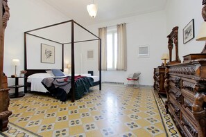 A four-poster bed with a white bedspread and a blanket, a wooden dresser with carved details, a chair, and a tiled floor with a geometric pattern. - Tiberina Spacious Apartment (Rome)