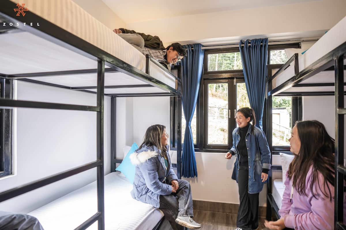 A dormitory room with bunk beds, a window with curtains, and a person standing and talking to another person sitting on a bed.
