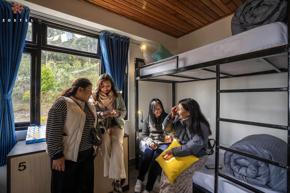 A dormitory room with bunk beds, a window with curtains, and a desk with a laptop.