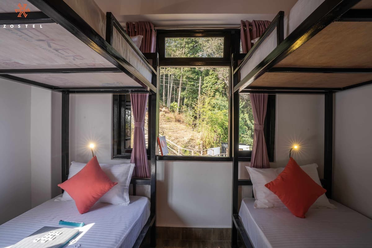 A bunk bed room with two beds, each with a pillow and a nightstand. A window provides a view of a forested area.