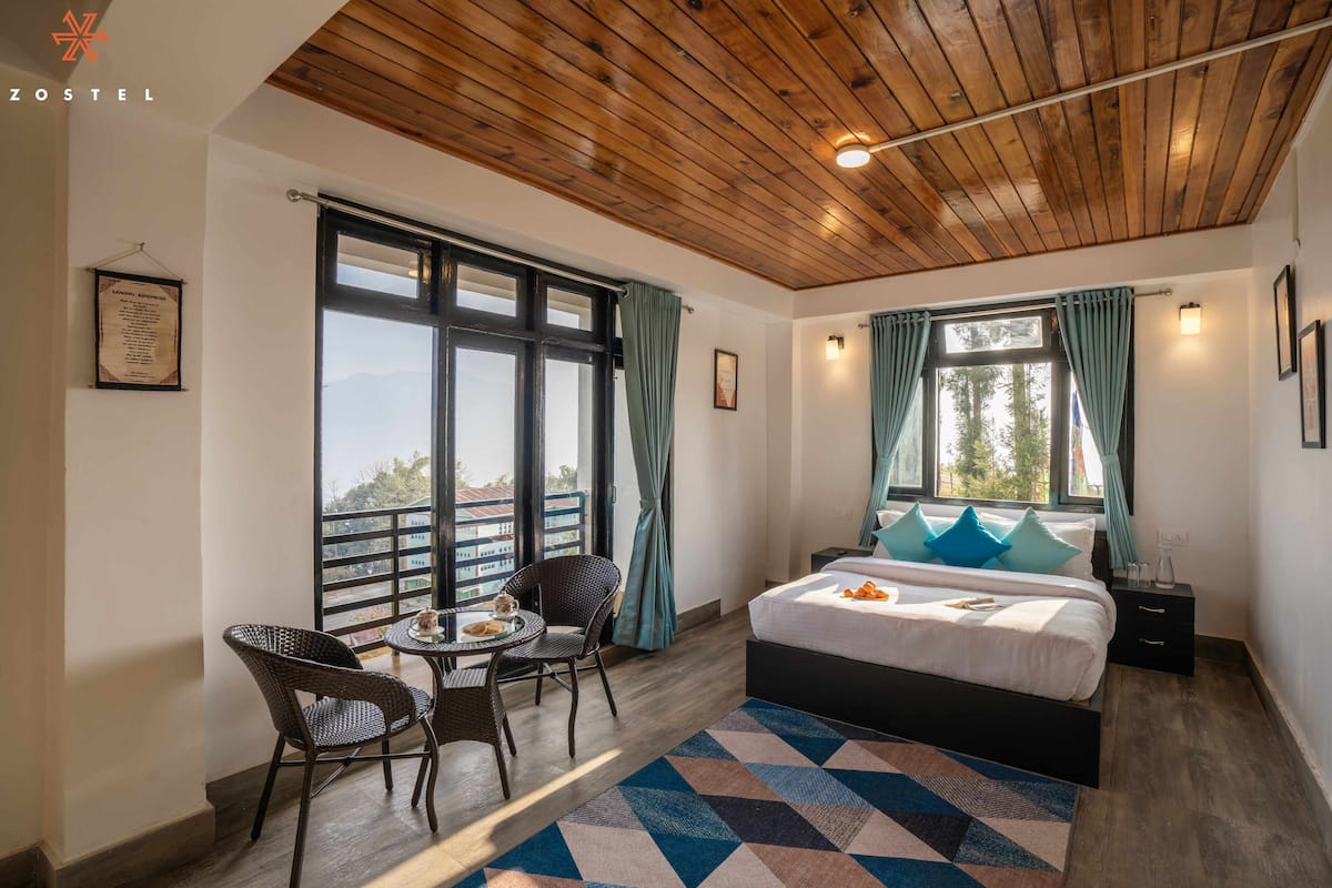 A bedroom with a bed, a small table with chairs, and large windows with curtains.