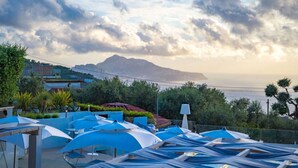 A coastal view with a mountain in the background, outdoor seating with umbrellas, and a clear sky. - Cosy apartment for 2 guests with A/C, WIFI, pool (Massa Lubrense)