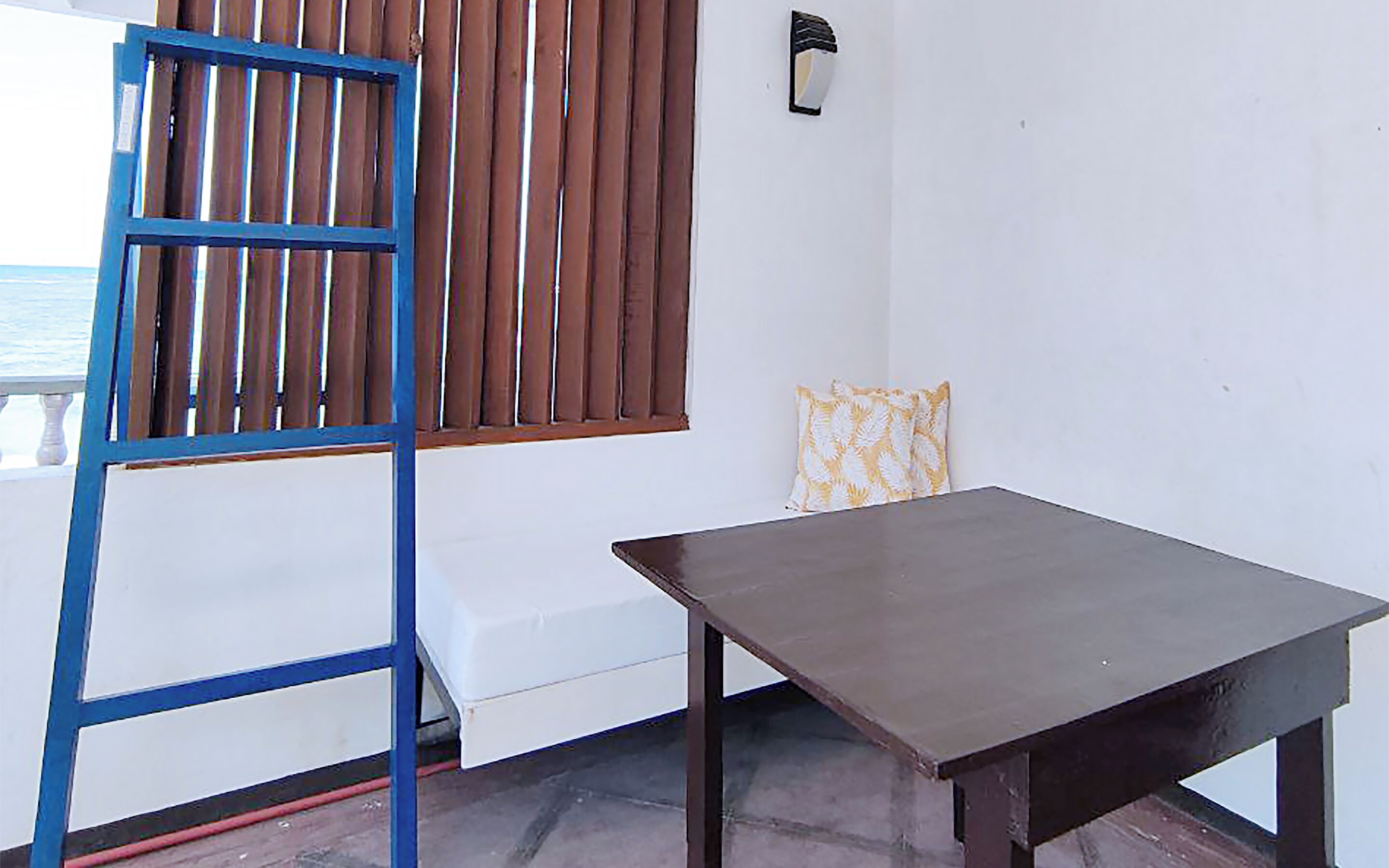Een kamer met een houten tafel, een blauwe metalen ladder, een raam met verticale zonwering en een gestreept kussen.