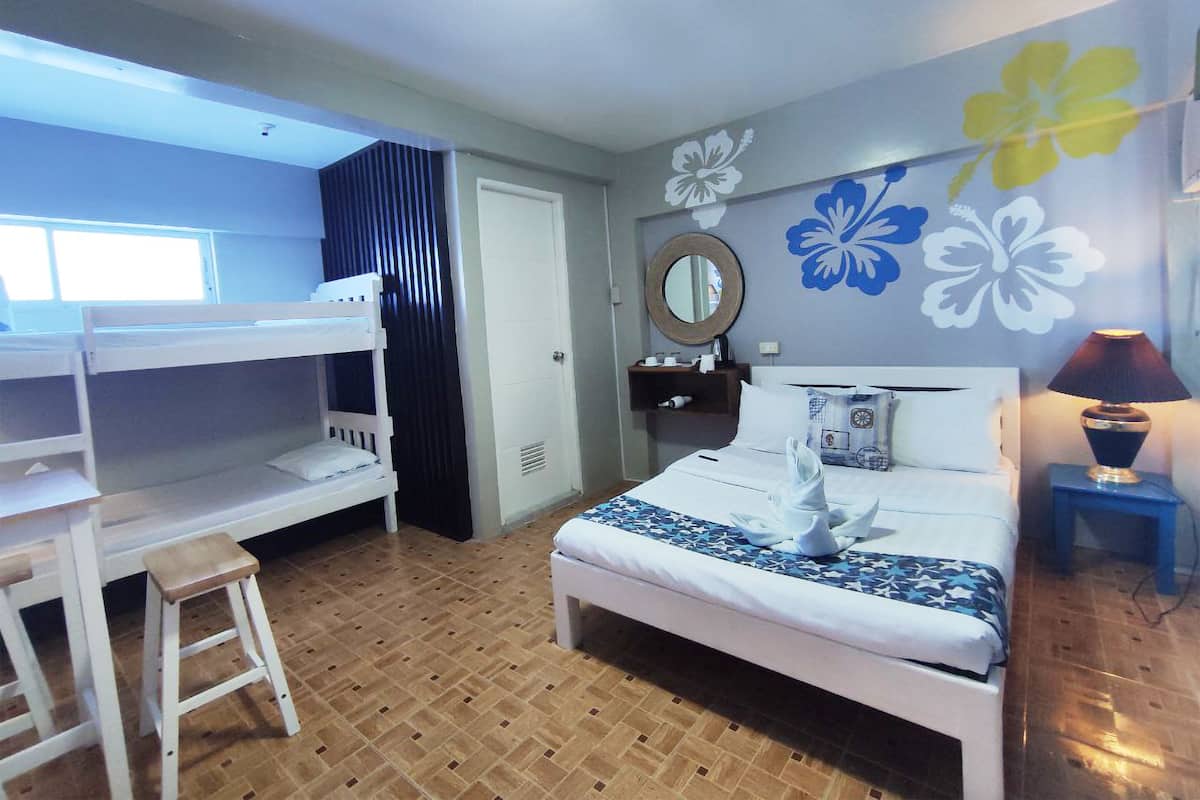 A hotel room with a bunk bed, a desk, a bedside table with a lamp, and a bed with a decorative pillow.