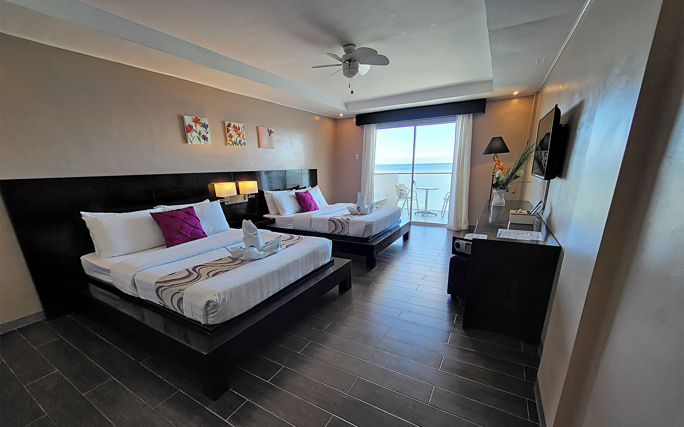 A hotel room with two beds, a desk, a ceiling fan, artwork on the wall, and a view of the ocean.
