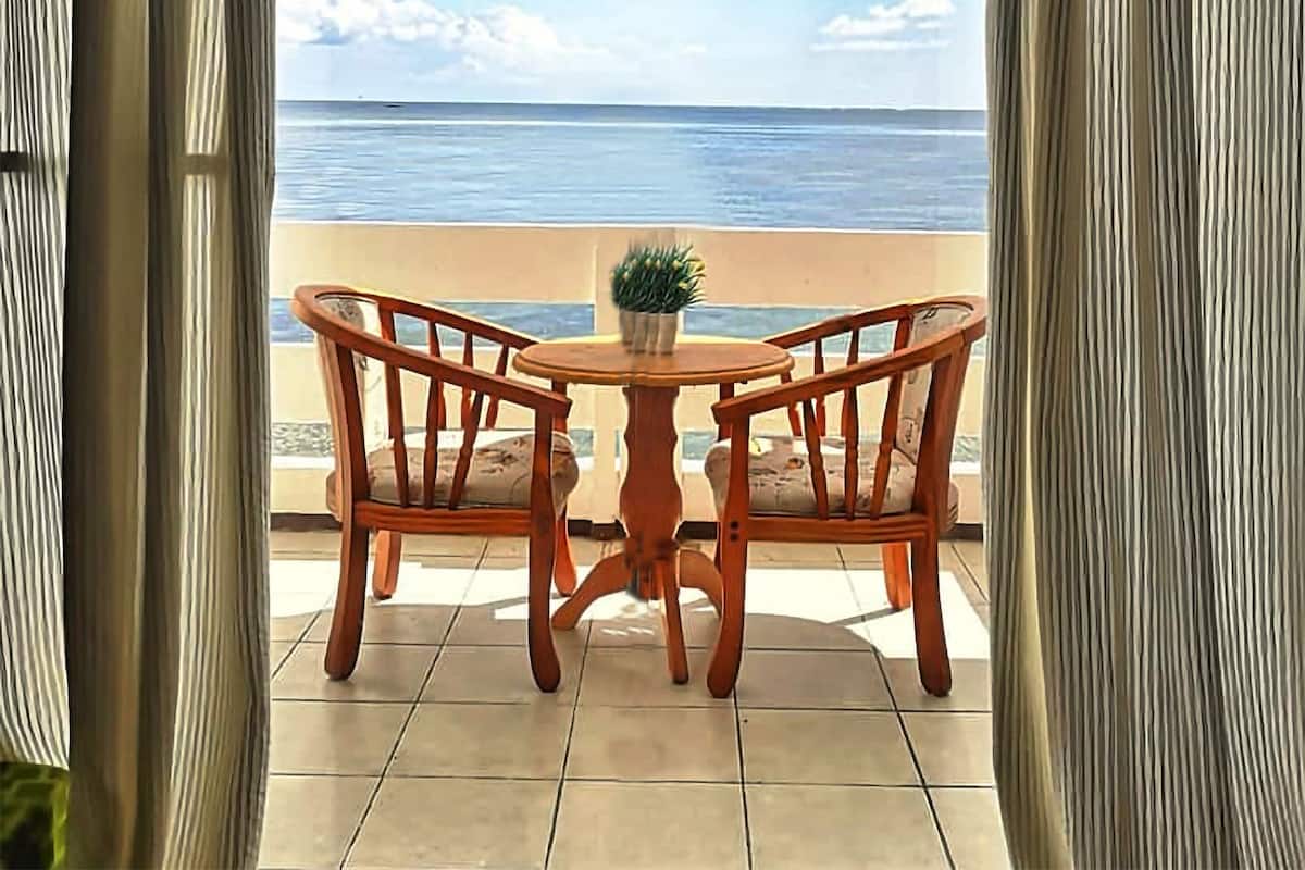 A view from a balcony with a wooden table and two chairs, overlooking a beach and the sea.
