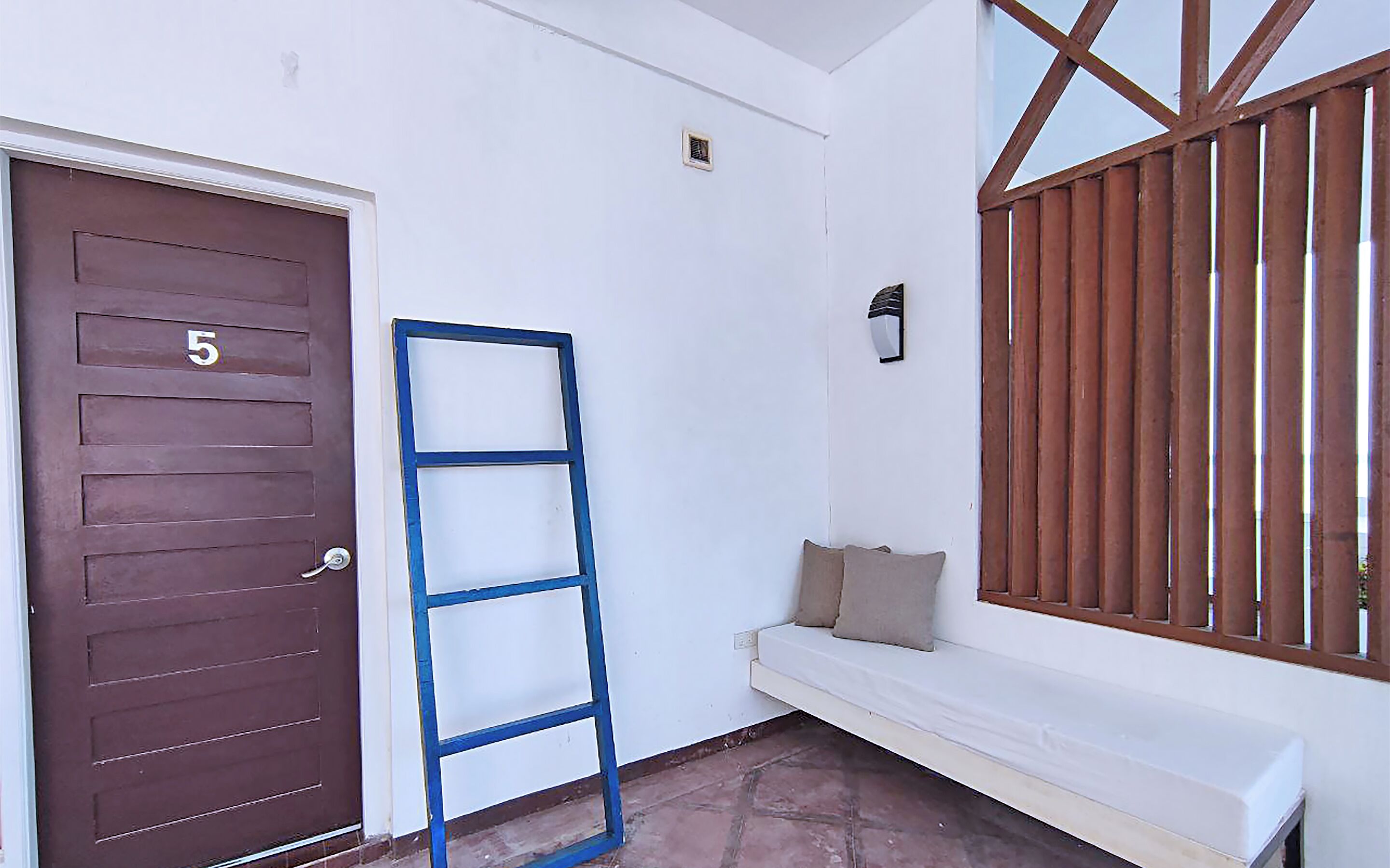 A room with a brown door marked with the number 5, a blue ladder leaning against the wall, a white bench with cushions, and a window with wooden bars.