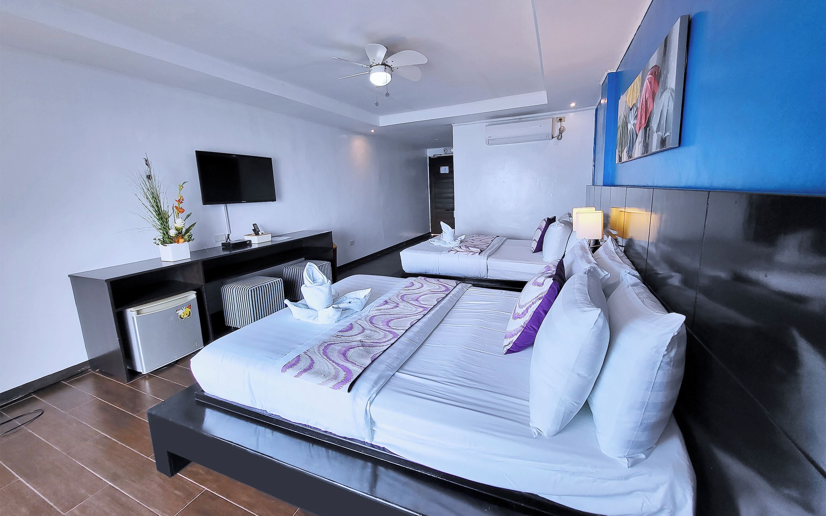 Une chambre d’hôtel moderne équipée de deux lits, d’un téléviseur à écran plat, d’un bureau avec un ordinateur et d’un ventilateur de plafond.