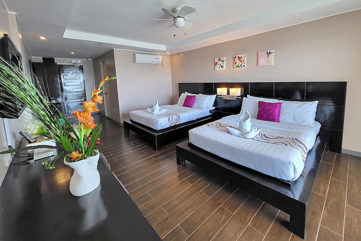 A hotel room with two beds, a ceiling fan, artwork on the wall, and a potted plant.