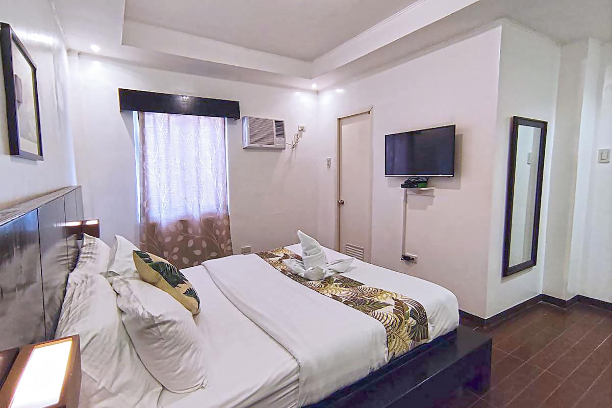 A hotel room with a bed, a TV mounted on the wall, an air conditioning unit, and a door.