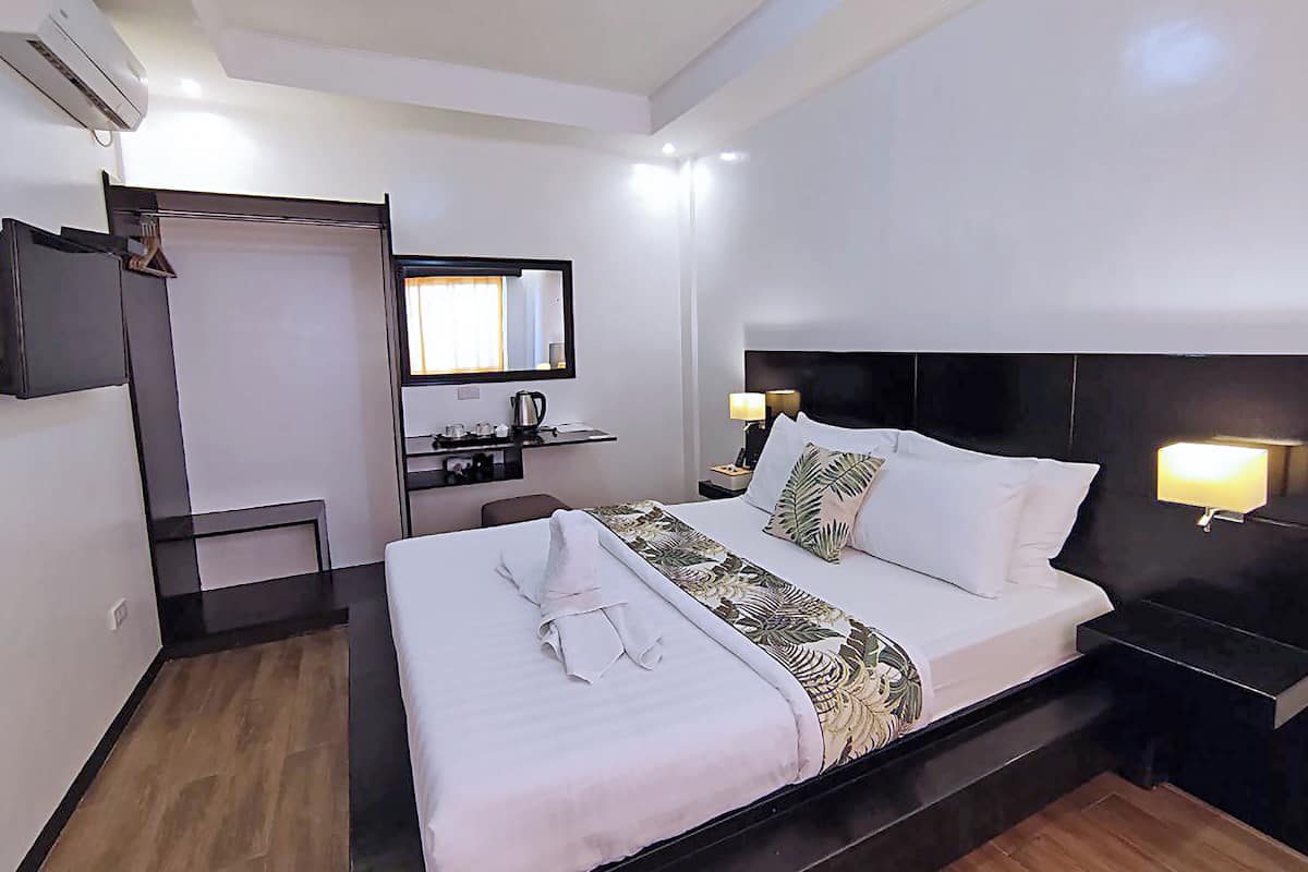 A hotel room with a bed, bedside table with lamps, a TV, a desk with a chair, and a small kitchenette.