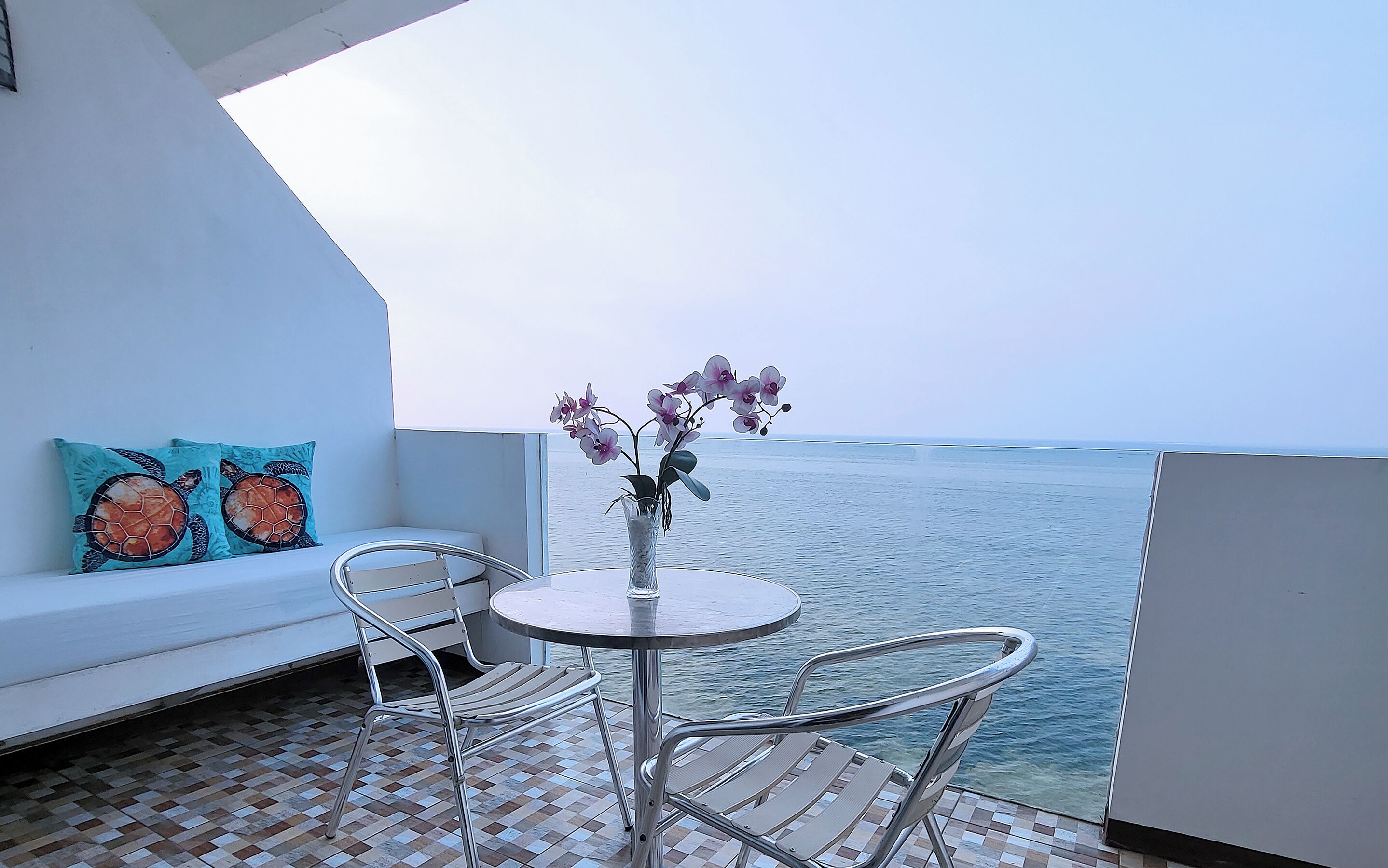 Un balcón con un sofá blanco, dos sillas de metal, una mesita redonda con un jarrón de flores y vista al mar.