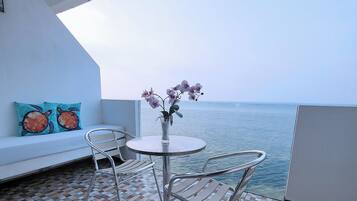 Un balcón con un sofá blanco, dos sillas de metal, una mesita redonda con un jarrón de flores y vista al mar.
