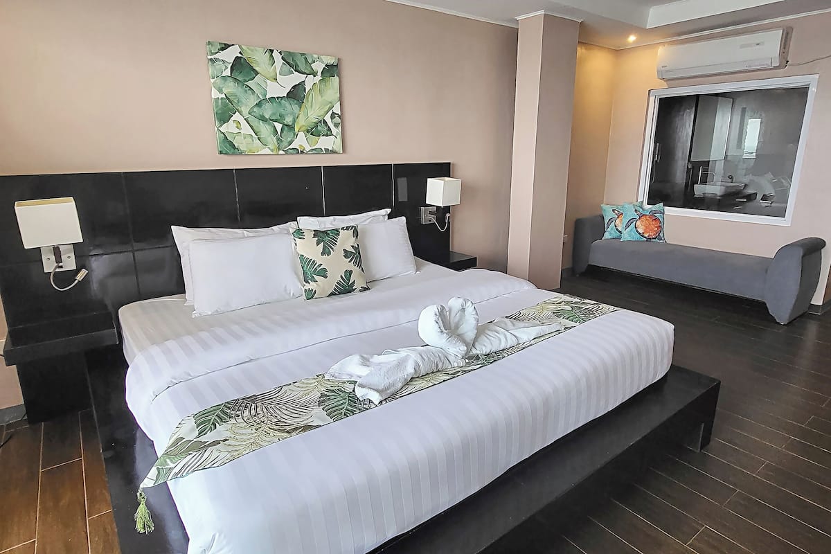A neatly made bed with white linens and a decorative pillow, a headboard with wall-mounted lamps, a large mirror, and a wall art piece above the bed.