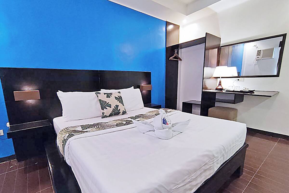 A hotel room with a large bed, a nightstand, a desk with a lamp, and a mirror.
