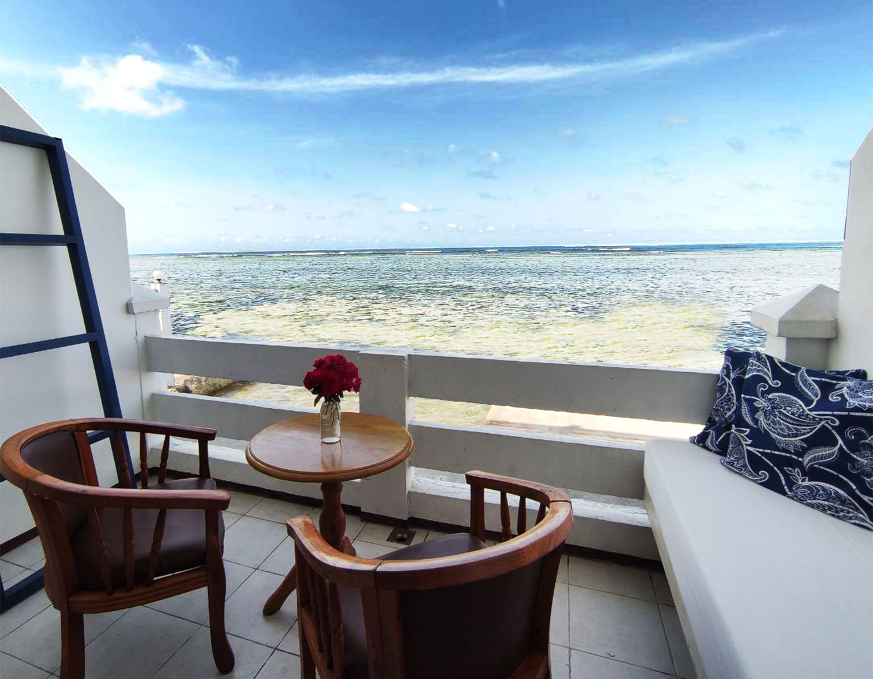 A balcony with two wooden chairs, a small round table with a vase of red flowers, and a sofa with a patterned cushion. The balcony overlooks a beach with the ocean in the background.