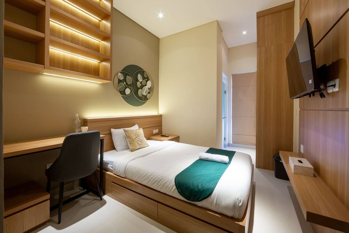 A modern hotel room with a wooden desk, a bed with a green throw, a wall-mounted TV, and a decorative wall piece.