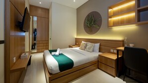 A modern hotel room with a wooden bed, a desk, a chair, a TV mounted on the wall, and a wall-mounted shelf with a plant. - Cove Tripuri House Bali (Tuban)