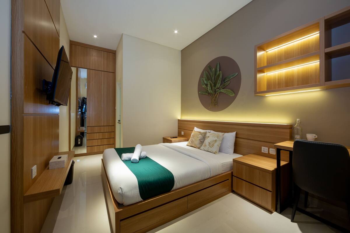 A modern hotel room with a wooden bed, a desk, a chair, a TV mounted on the wall, and a wall-mounted shelf with a plant.